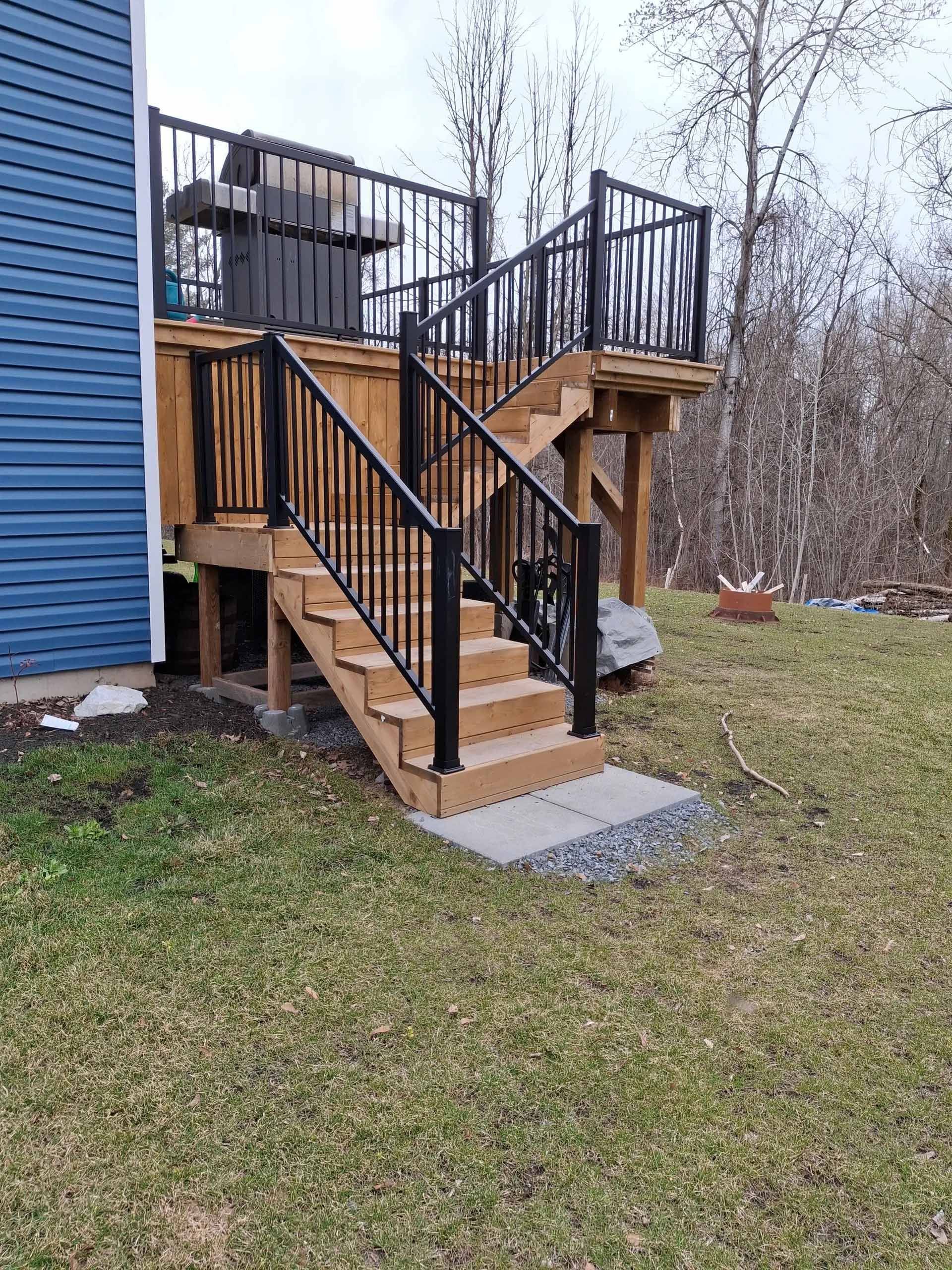 Deck with black railings and steps leading to a yard, set against a blue-sided house and trees.