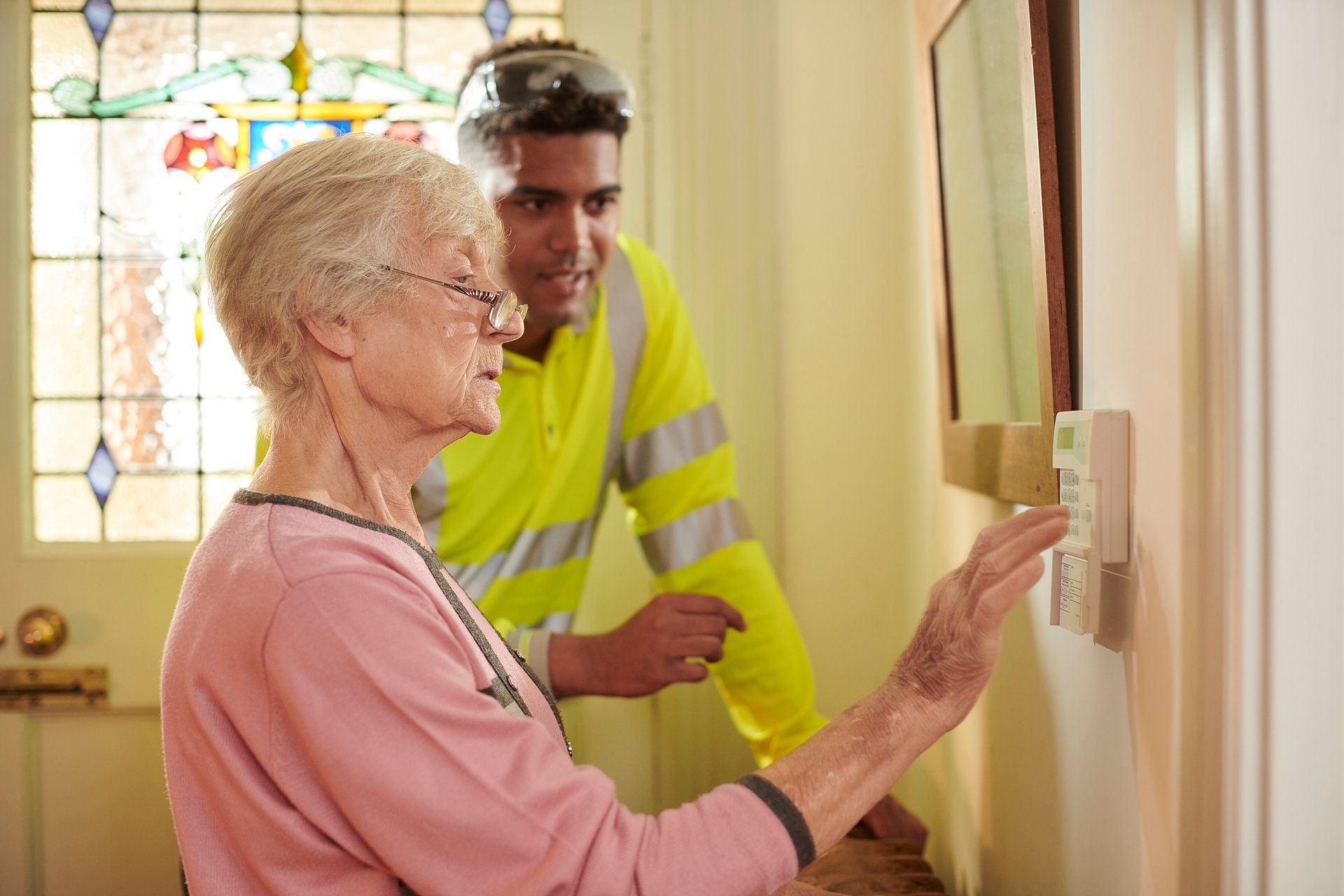 An elderly woman is adjusting a thermostat while a man looks on.