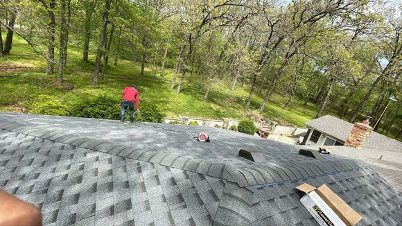 Person on a roof installing shingles, surrounded by trees.