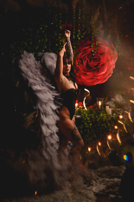 A woman in a black bodysuit with angel wings is standing in front of a large red rose.