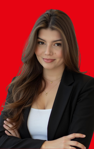 Woman with long brown hair, wearing a black blazer, white top, and a necklace, arms crossed, smiling against a red backdrop.