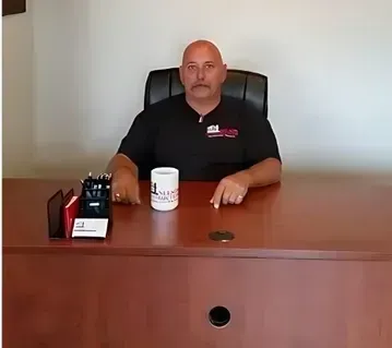 Man sitting at a desk with a coffee mug and office supplies. Wearing a black shirt, in an office.