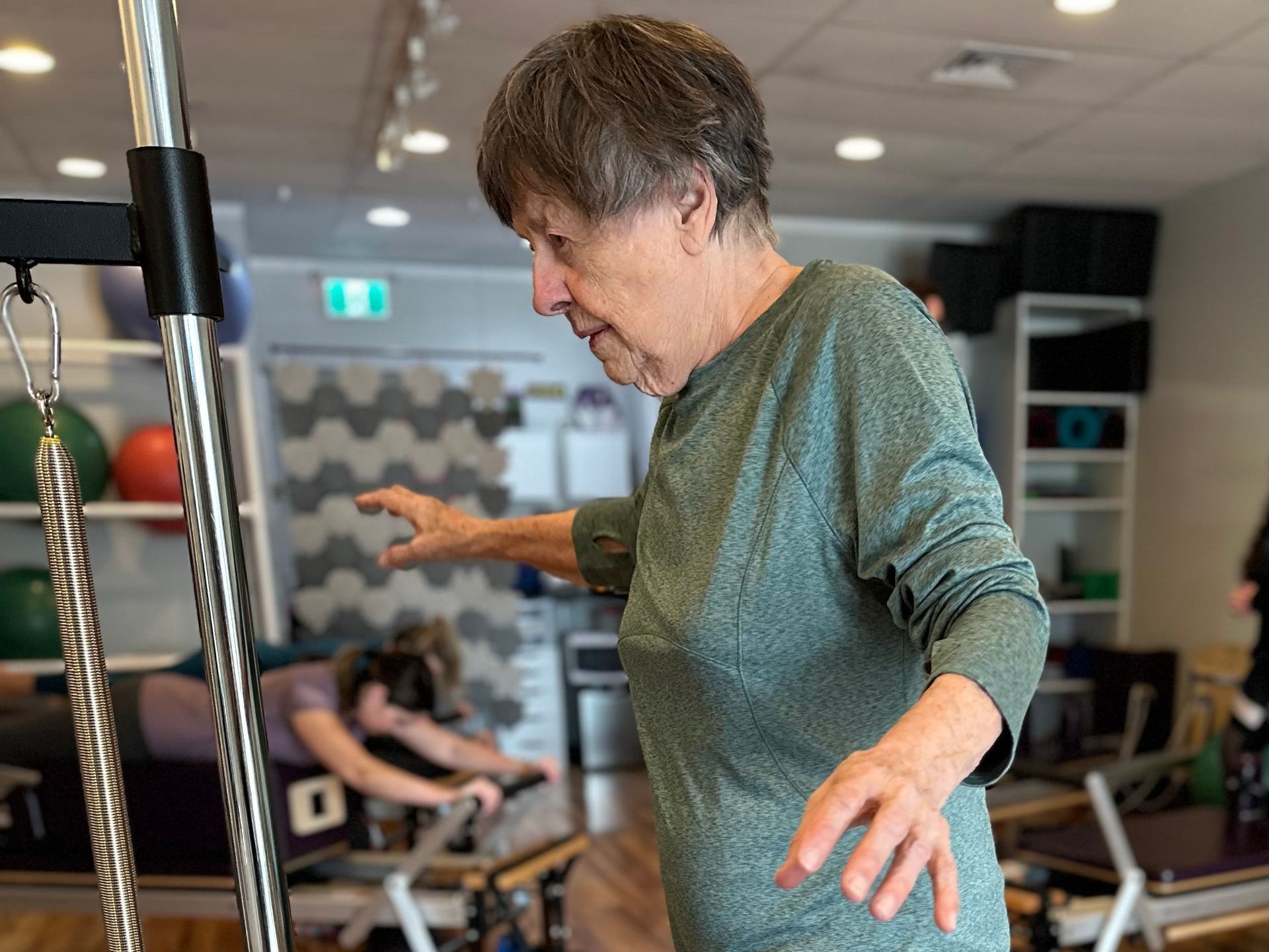 An elderly woman is standing on a pilates machine in a gym.