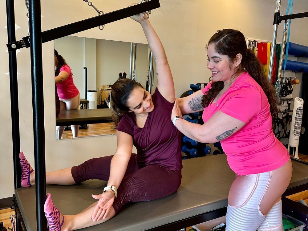 A woman is helping another woman stretch her legs on a pilates machine.