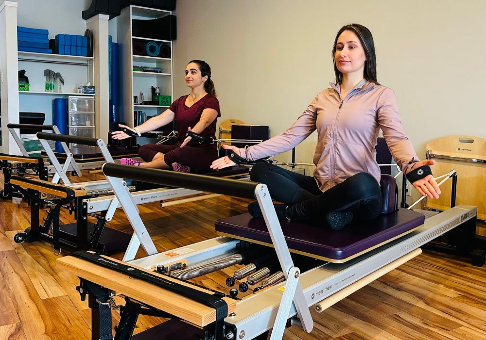 Two women are sitting on pilates machines in a gym.