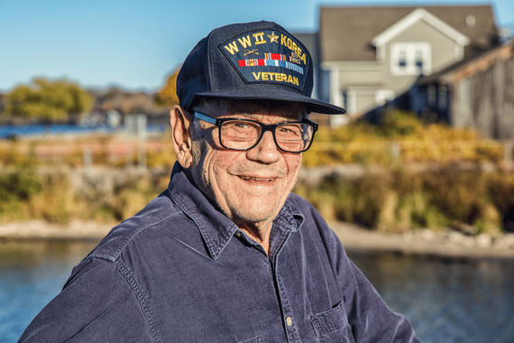 Elderly WWII veteran wearing a cap and glasses smiles outdoors near water and buildings.