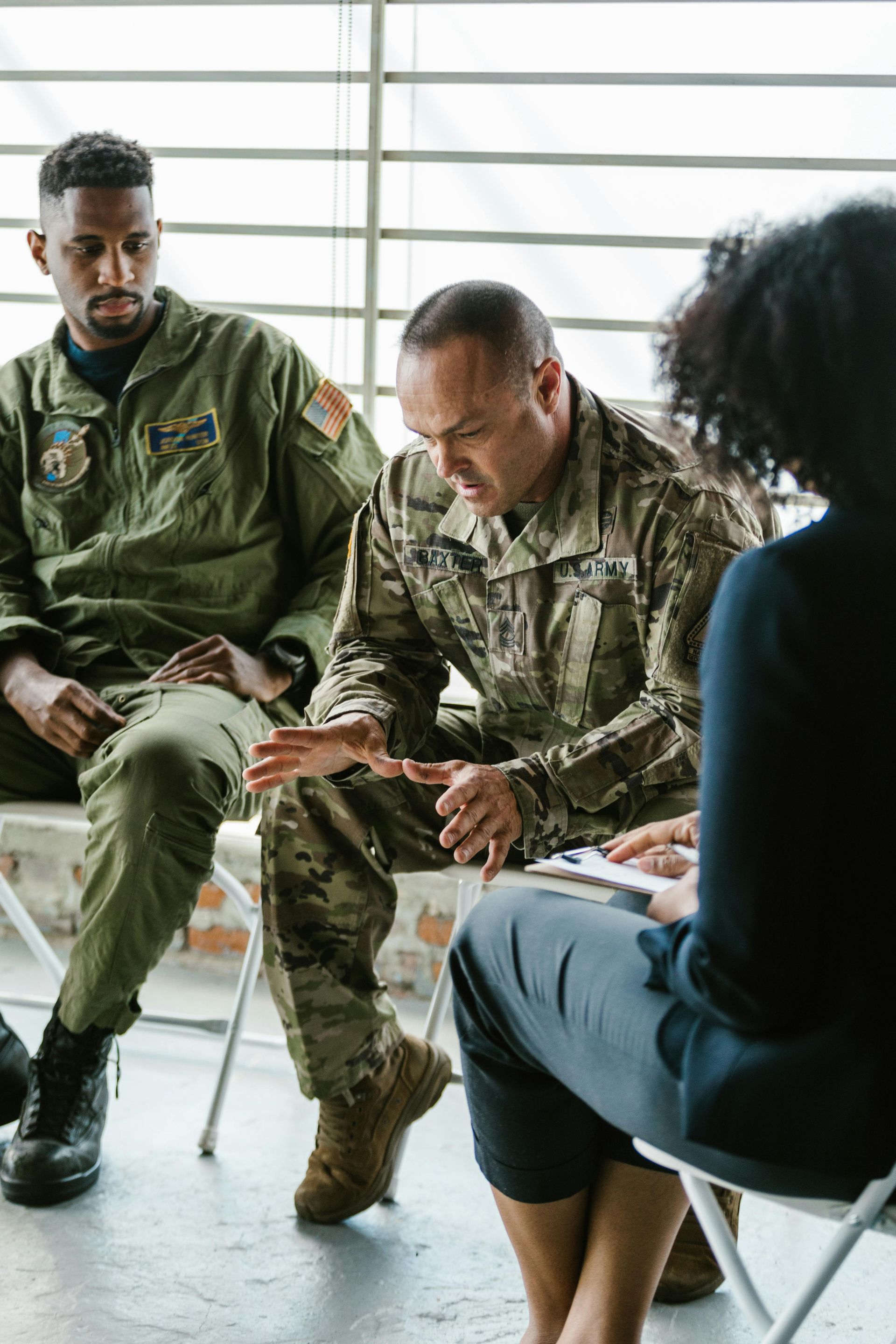Three people in a circle. Two military men, one in a flight suit, another in camo, seated near a woman. They are having a discussion.