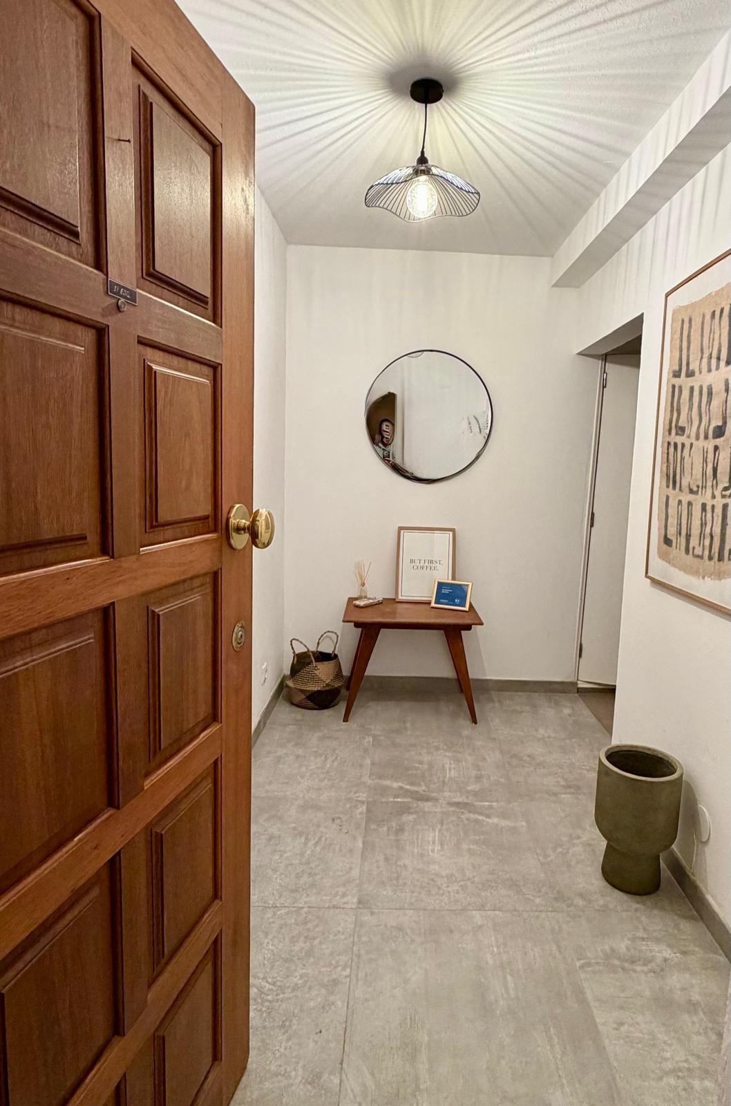 Gang met witte muren, een ronde spiegel, kunstwerken, een klein tafeltje en open deuren.