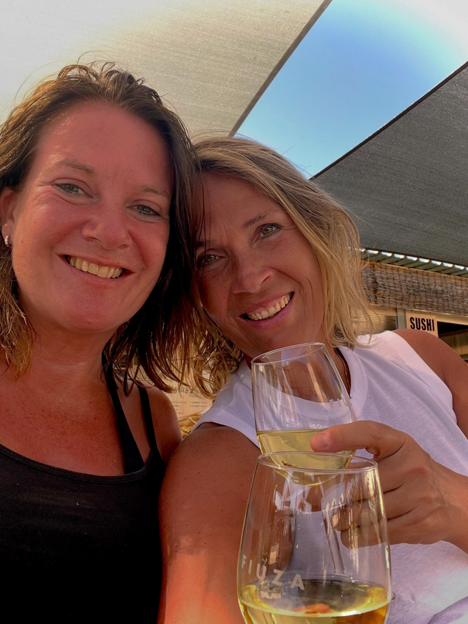 Twee lachende vrouwen, waarvan één met een glas witte wijn, in de halfschaduw op een zonnige dag.