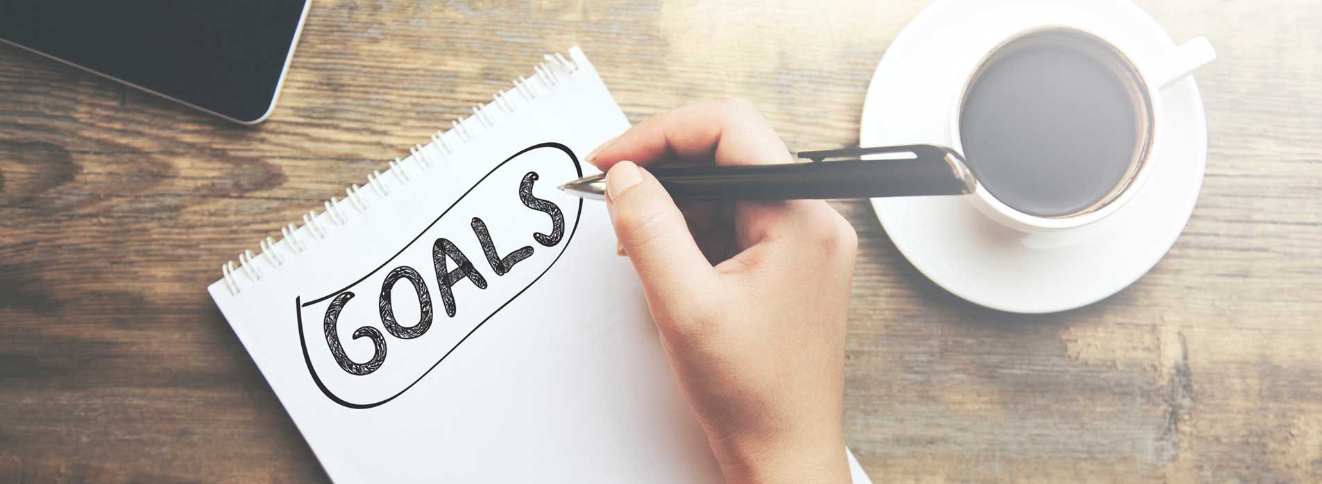 A person is writing goals on a piece of paper next to a cup of coffee.