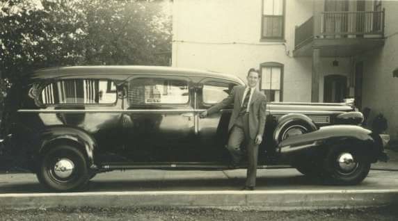 Man in a suit stands next to a large, dark car. Building in the background.