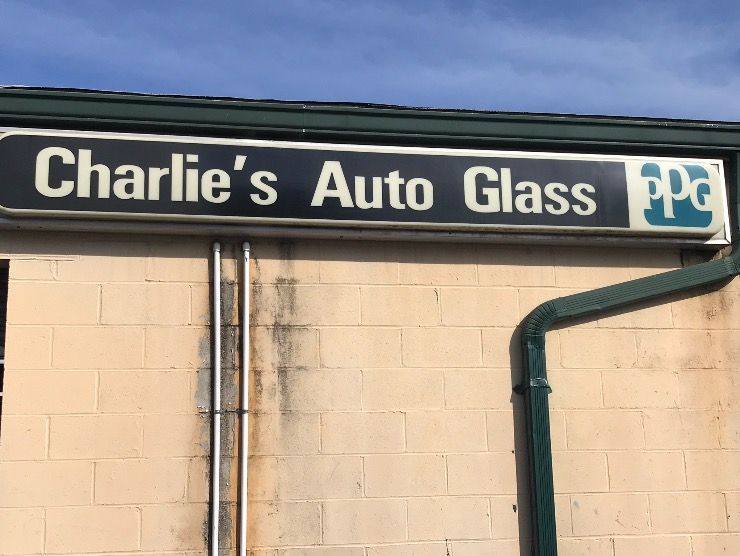 A sign for Charlie’s Auto Glass with a blue PPG logo on a beige brick building with a green trim and downspout.