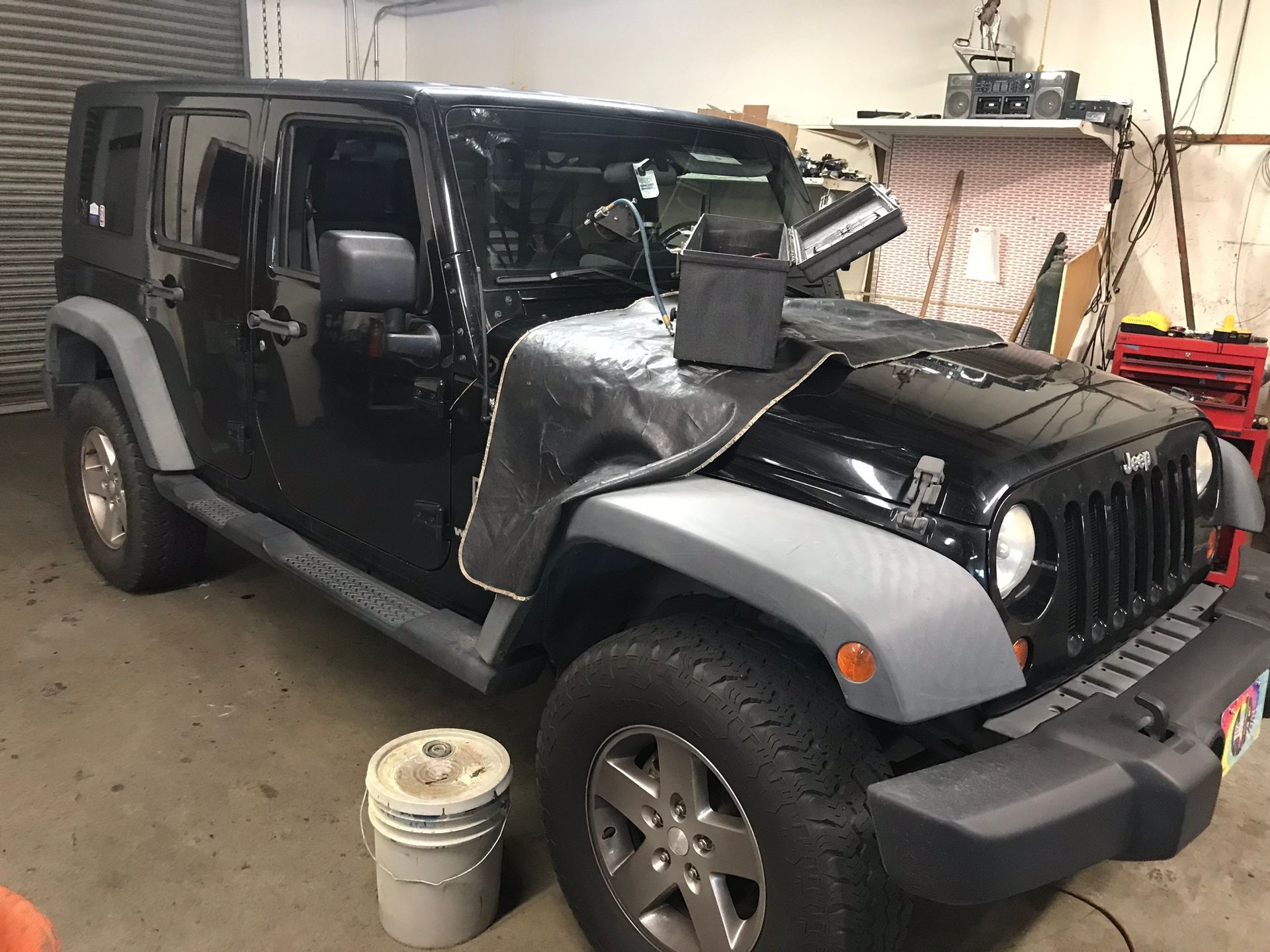 A black Jeep with grey fenders sits inside a garage, with a protective cover draped over the hood.