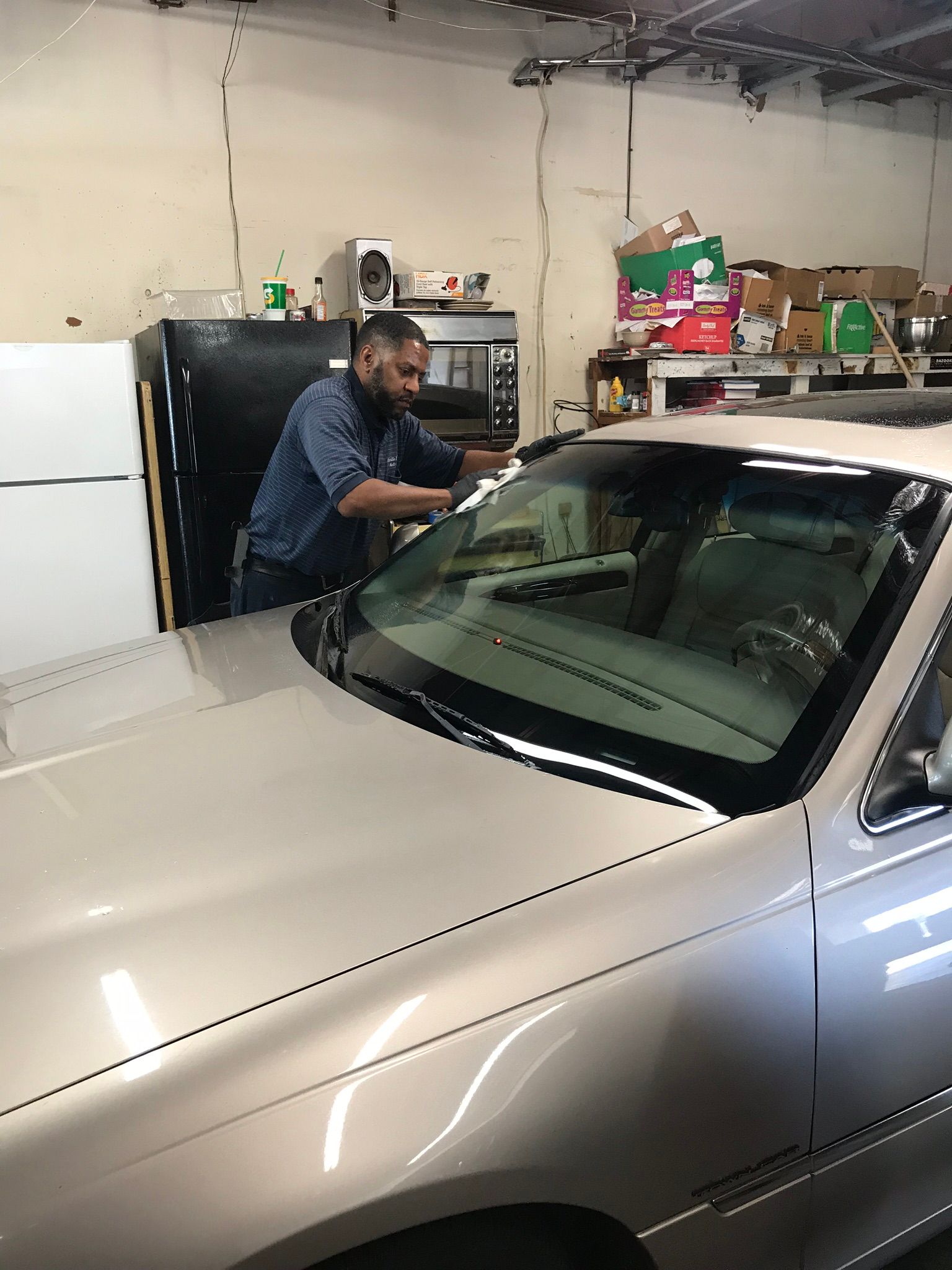 A person in a blue shirt works on the windshield of a beige sedan parked inside a garage.