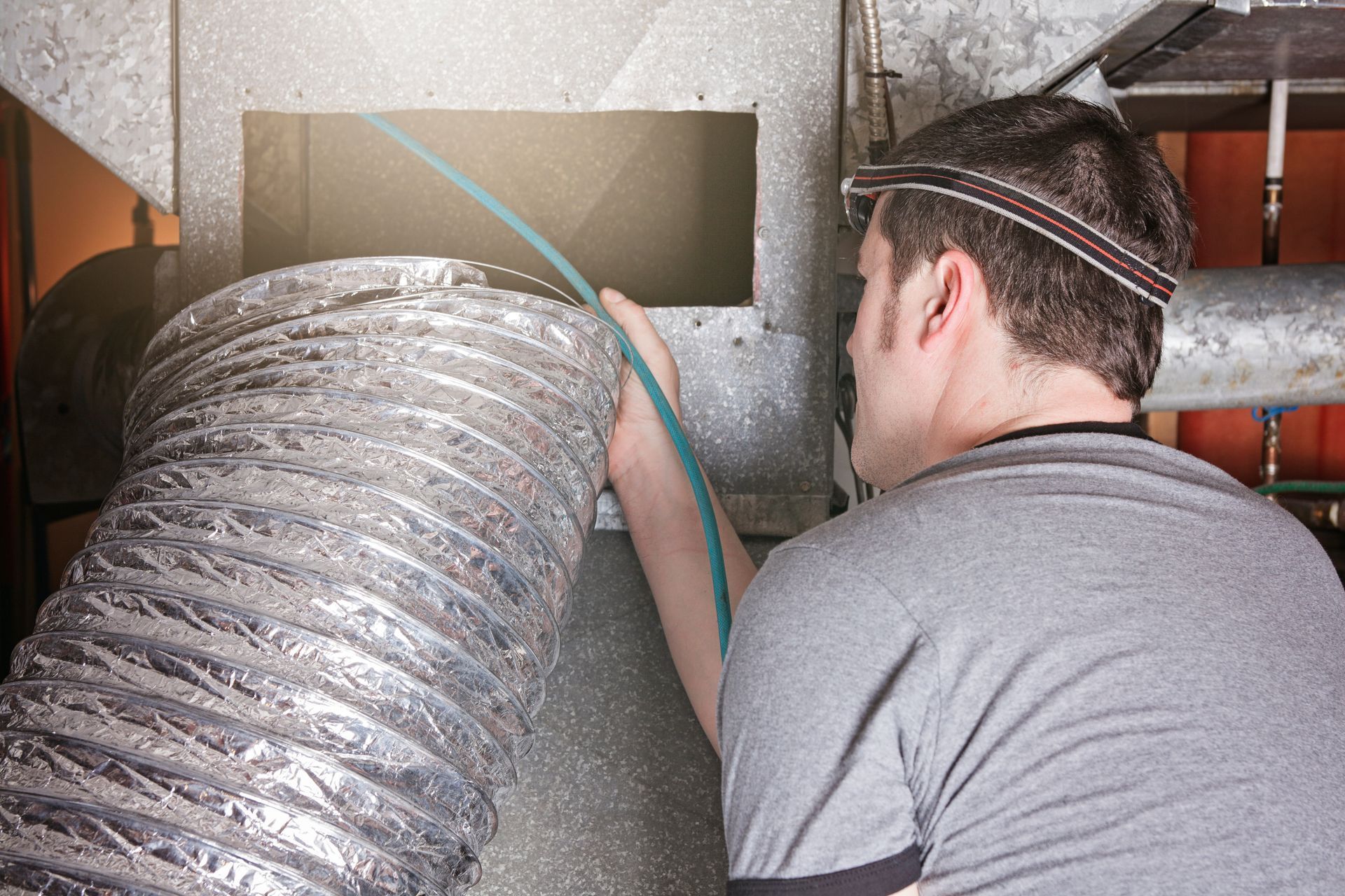 Man in blue overalls on a ladder, installing HVAC ductwork in a hallway.