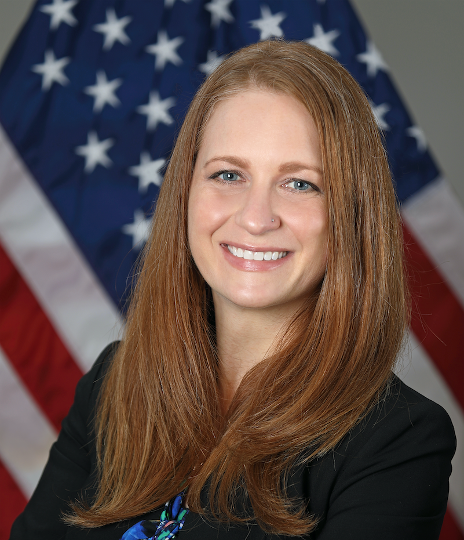 Image of Tara Schwetz (white woman with red hair, blue eyes) in a black blazer in front of an American flag
