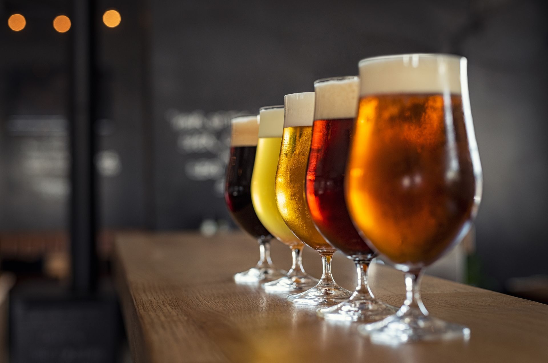 A row of glasses filled with different types of beer are lined up on a bar.