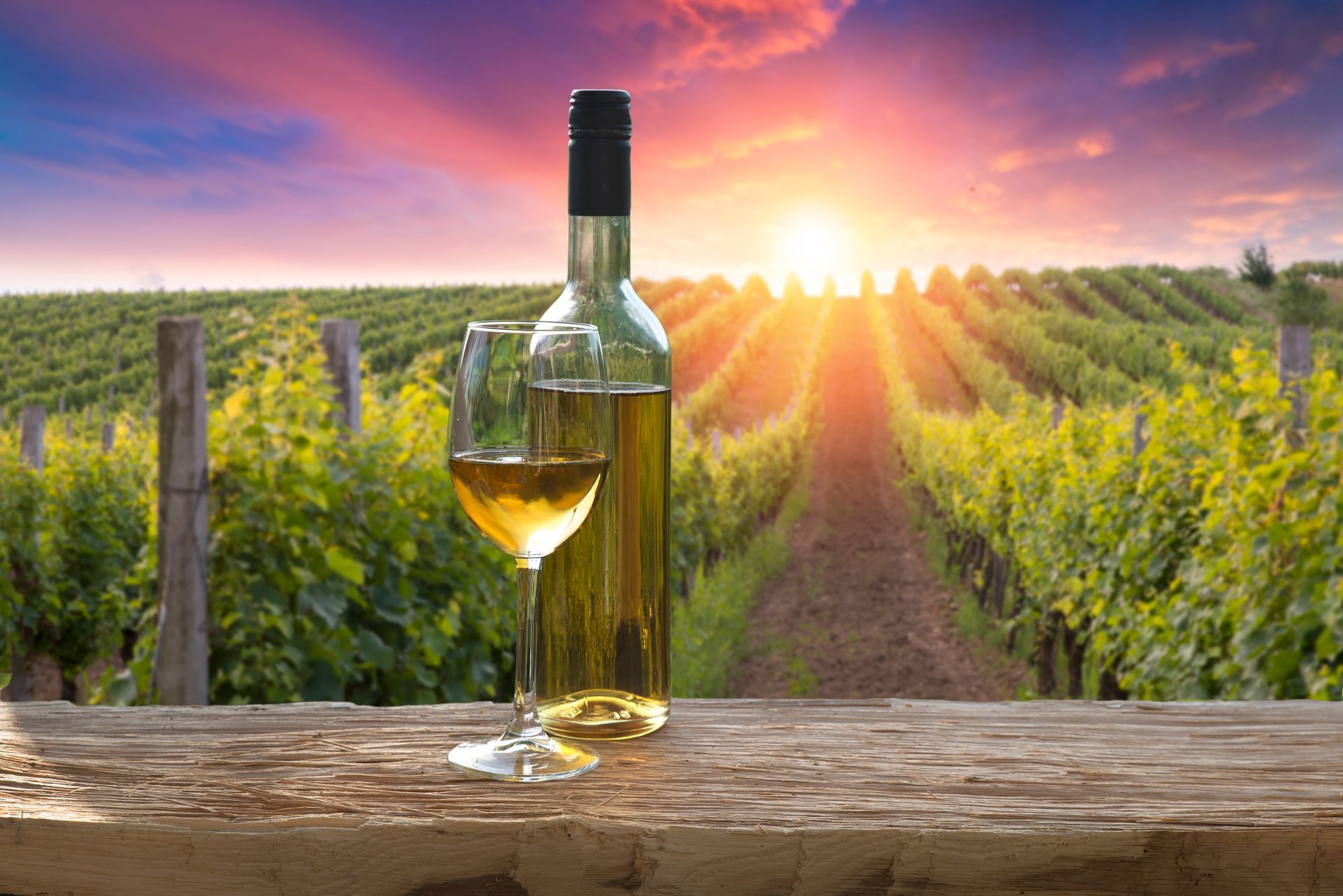 A bottle of wine and a glass of wine are on a wooden table in front of a vineyard.