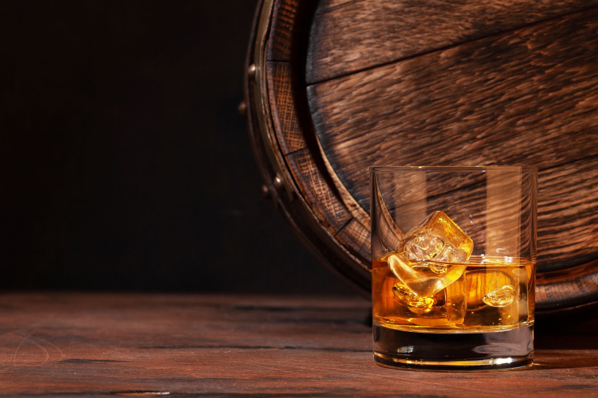 A glass of whiskey with ice cubes next to a wooden barrel.