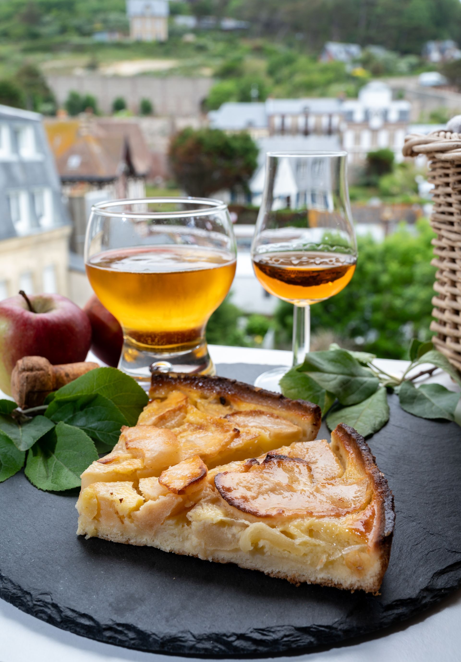 A slice of apple pie is on a cutting board next to two glasses of wine.