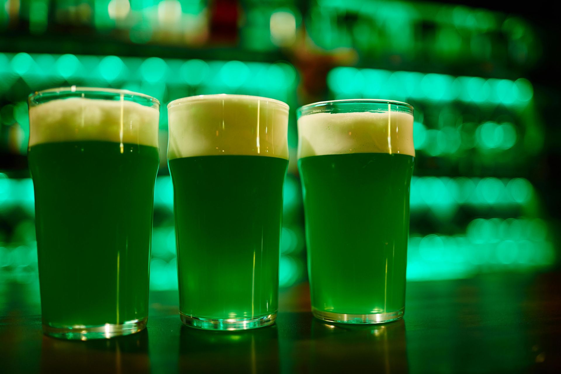 Three glasses of green beer are sitting on a bar counter.