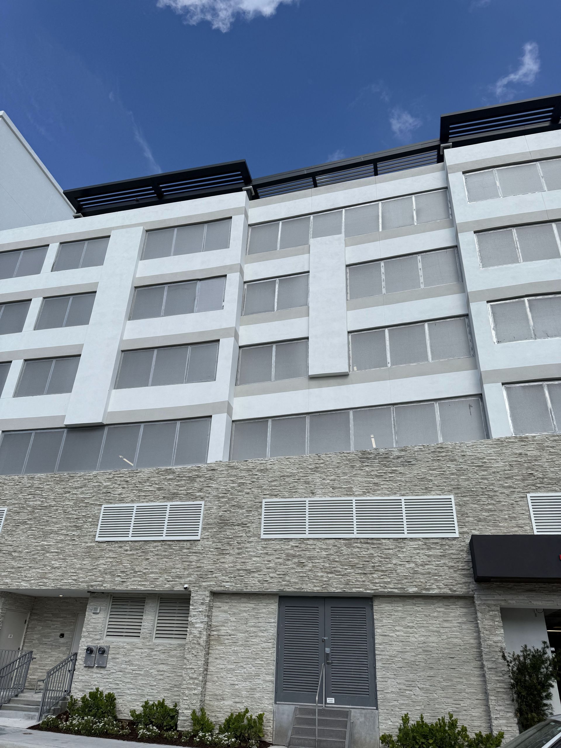 Modern white building with stone base, many windows, and a dark roof against a blue sky.