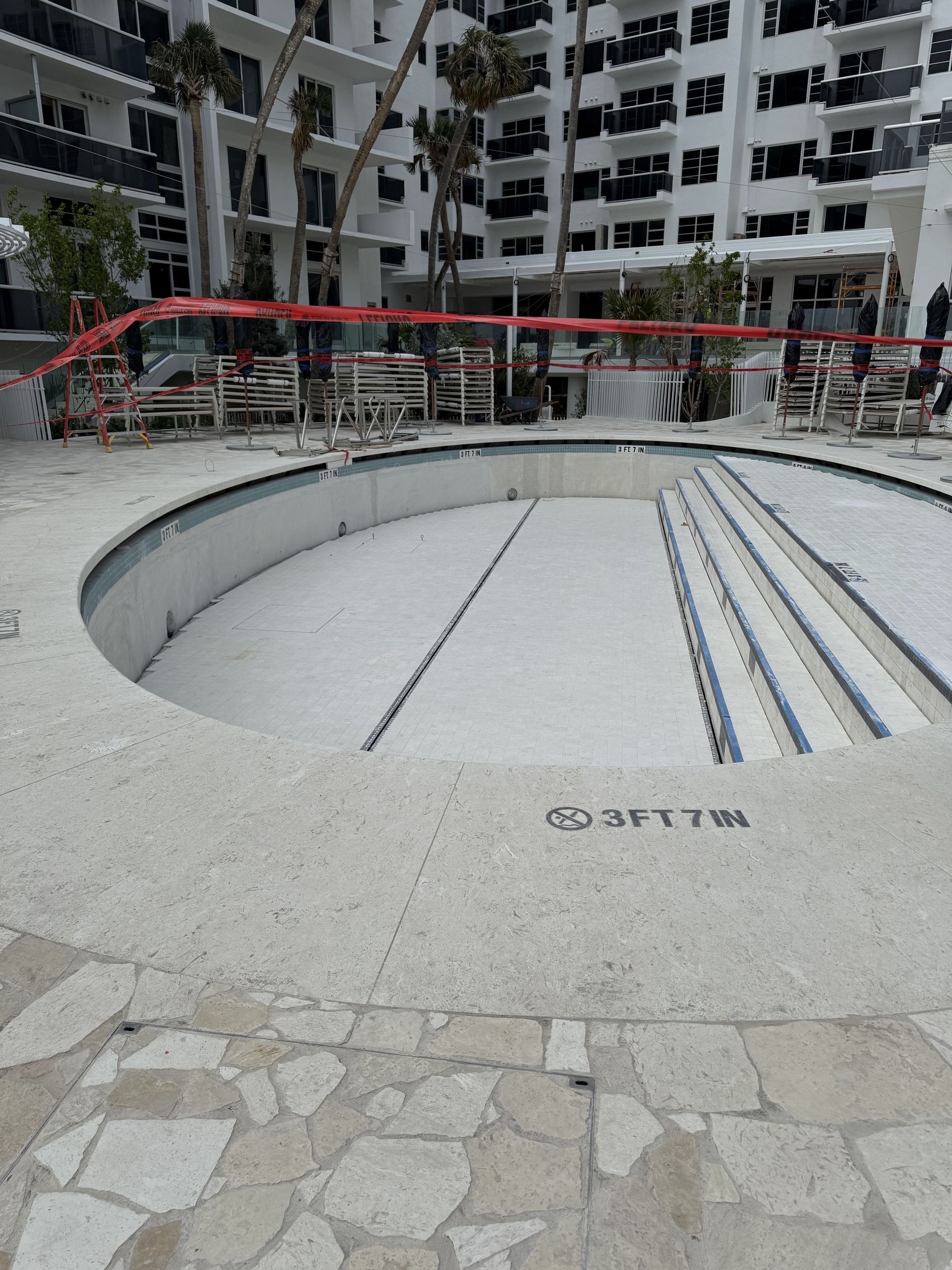 Empty pool under construction, with a white exterior, and a building in the background. Red tape blocks access.