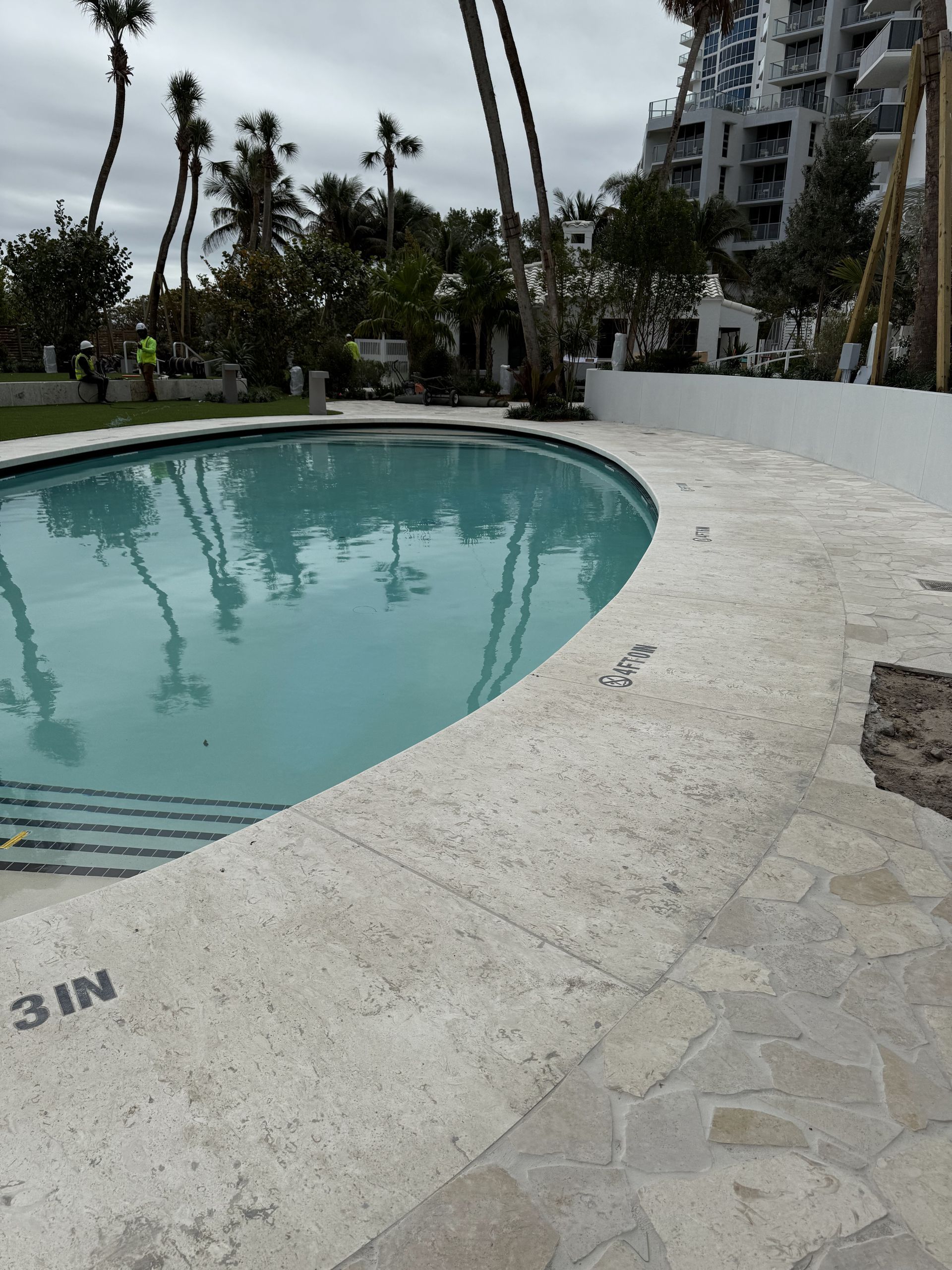 Pool with light-colored stone surround. Palm trees and a building are in the background. The water is still.