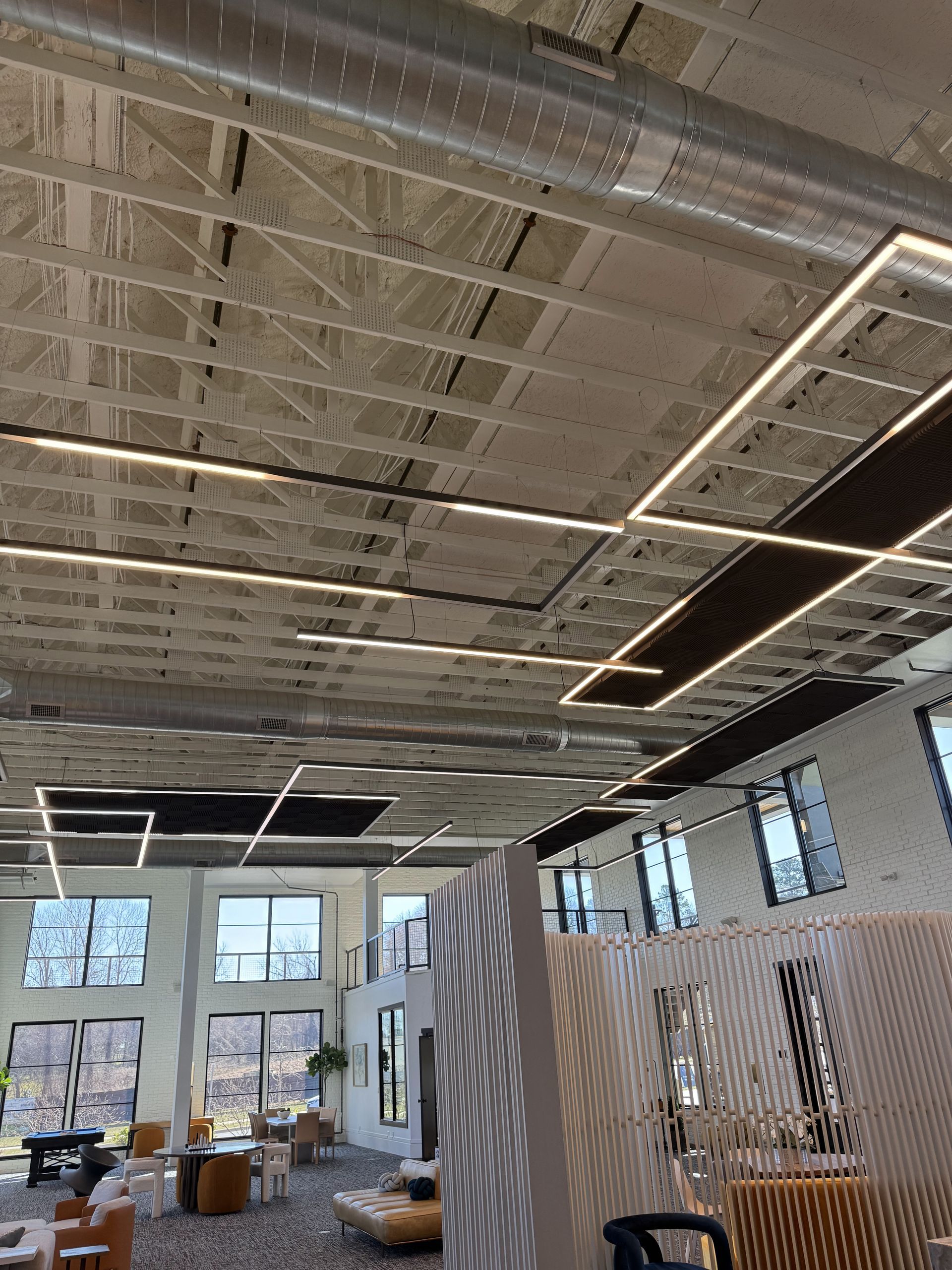 High-ceilinged room with exposed concrete ceiling, linear lighting, windows, and partial view of furniture.