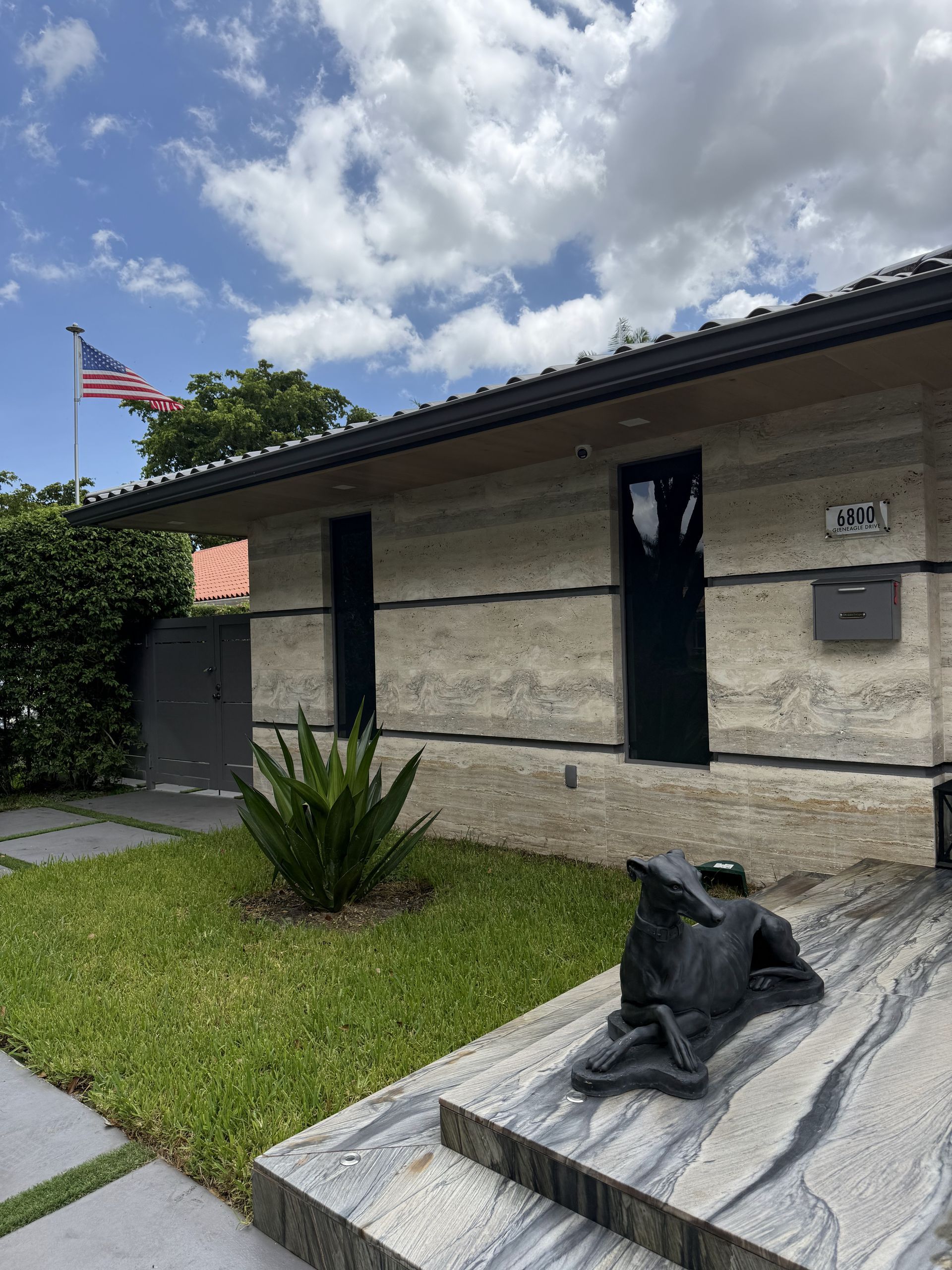 A light-colored stone house with an American flag, black door, mailbox, and a statue of a dog.