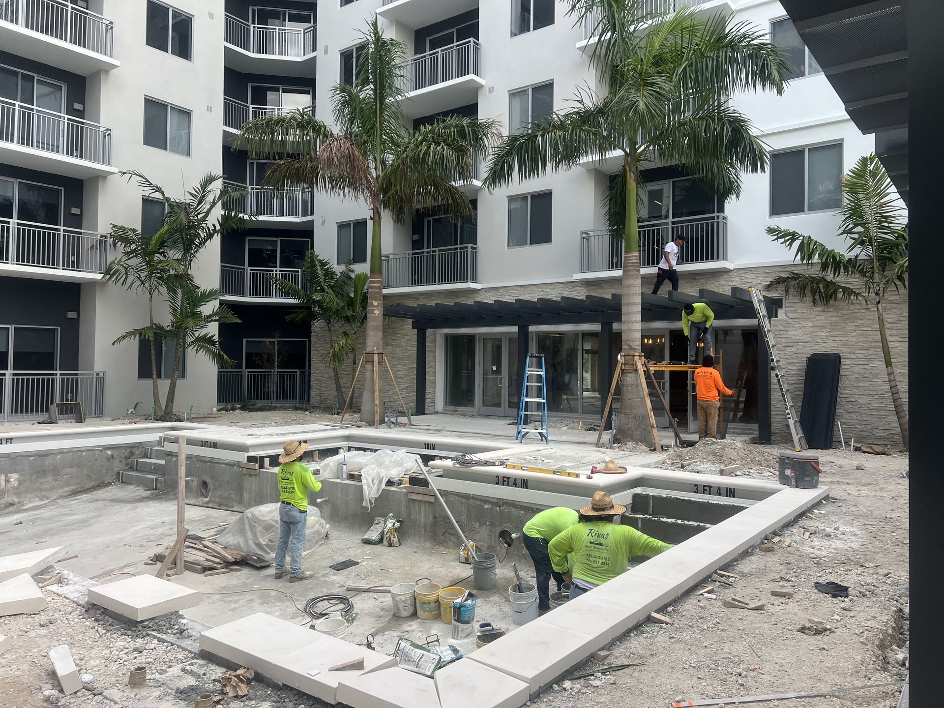 Construction site: workers near a pool under renovation, with a multi-story white building in the background.