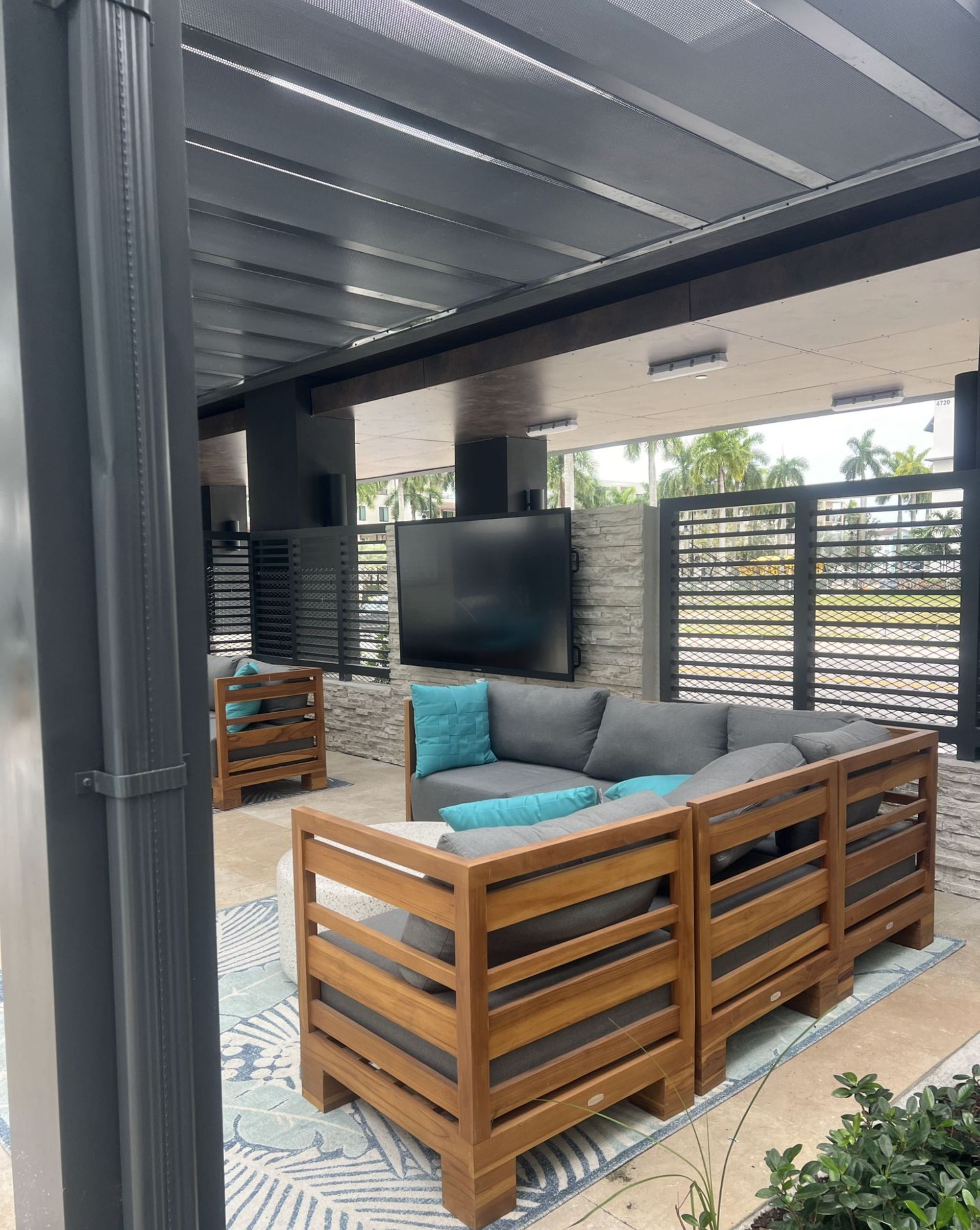 Outdoor seating area with gray sectional, TV, wood furniture, and overhead slats.