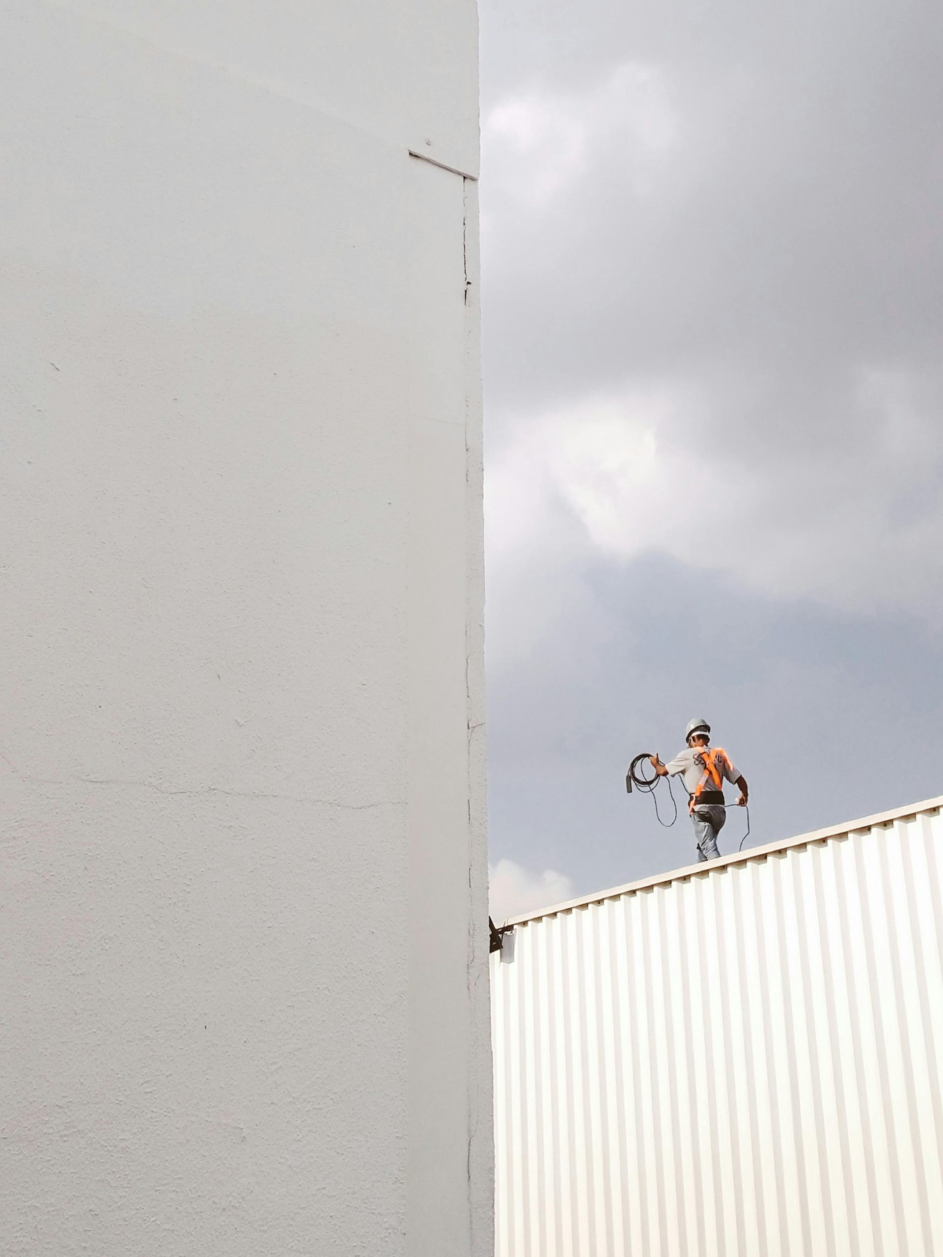 Man on a corrugated metal roof, wielding a tool. Building corner on the left, cloudy sky.