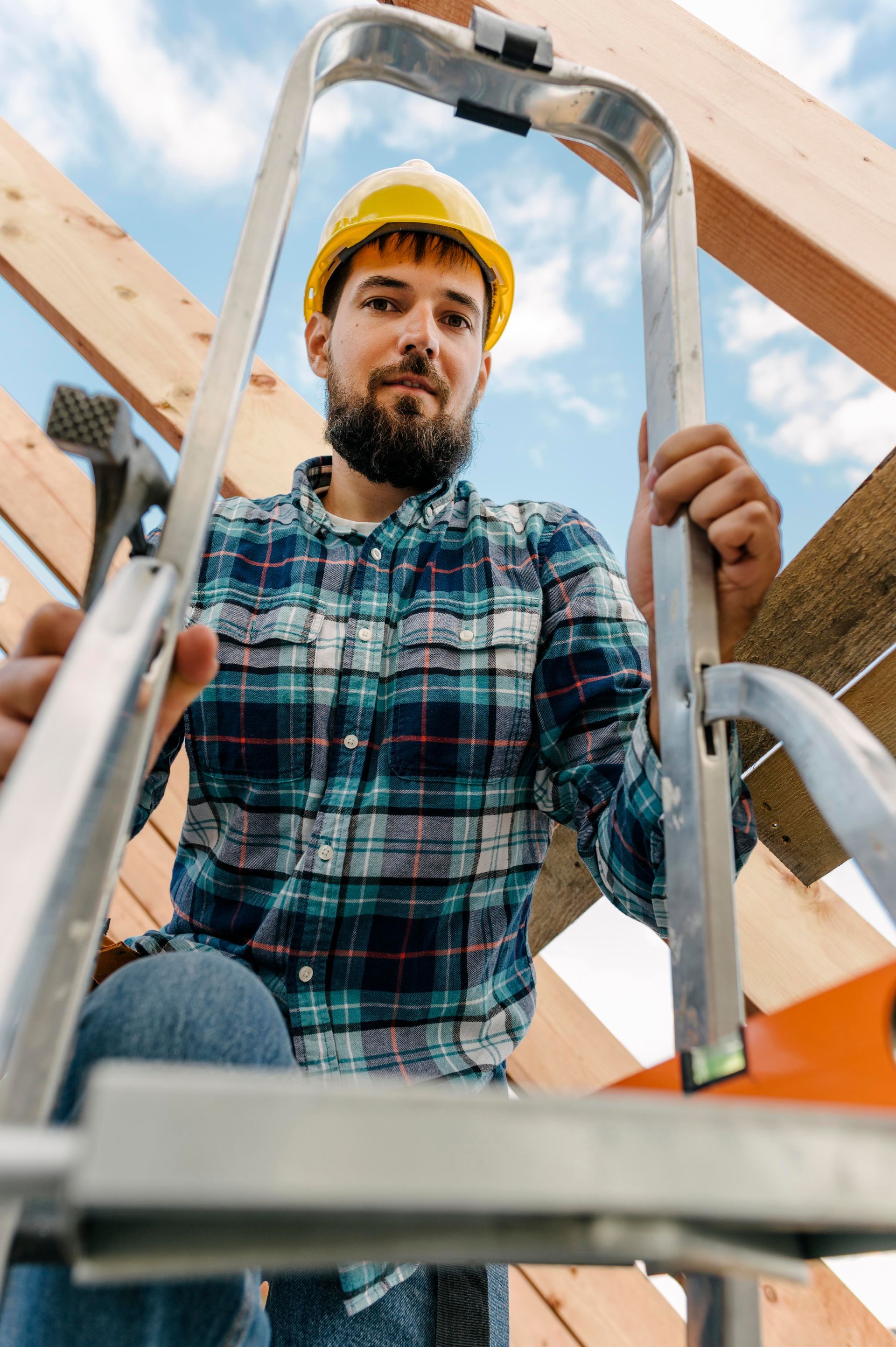 Construction worker on ladder, wearing yellow hard hat and plaid shirt, amidst roof framework, looking at camera.