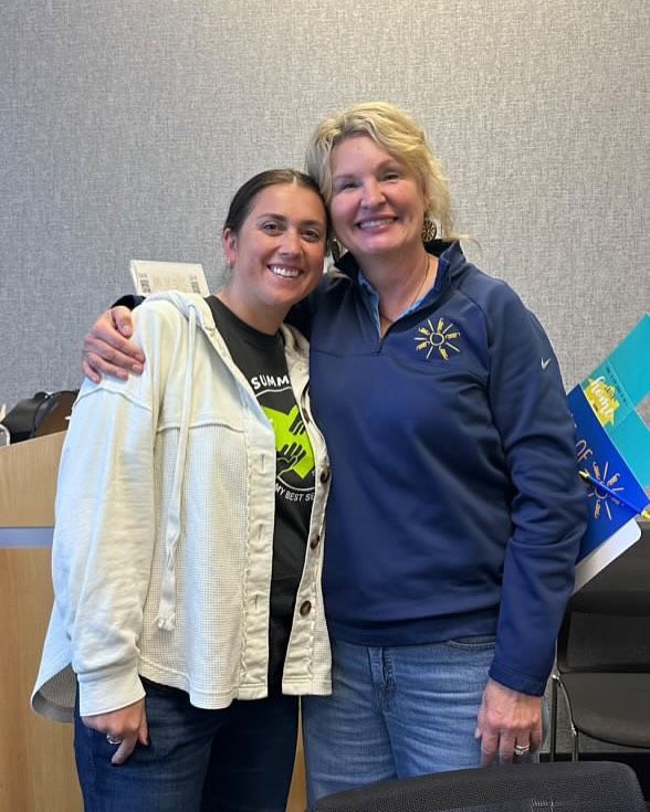 Two women smile while embracing in an office setting. The woman on the left wears a light jacket, and the woman on the right has a blue jacket.