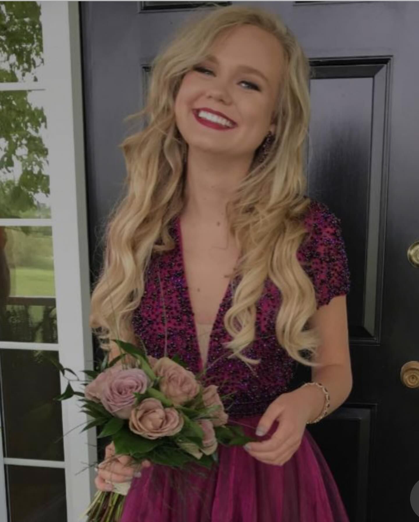 Blonde woman in a magenta dress smiles, holding flowers. She stands outside a dark door with a white trim.
