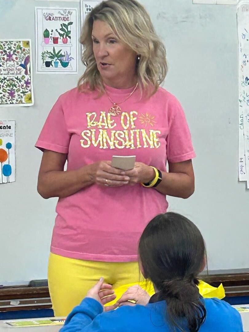 A woman in a pink shirt and yellow pants speaks to a student in a classroom. She holds a small item.
