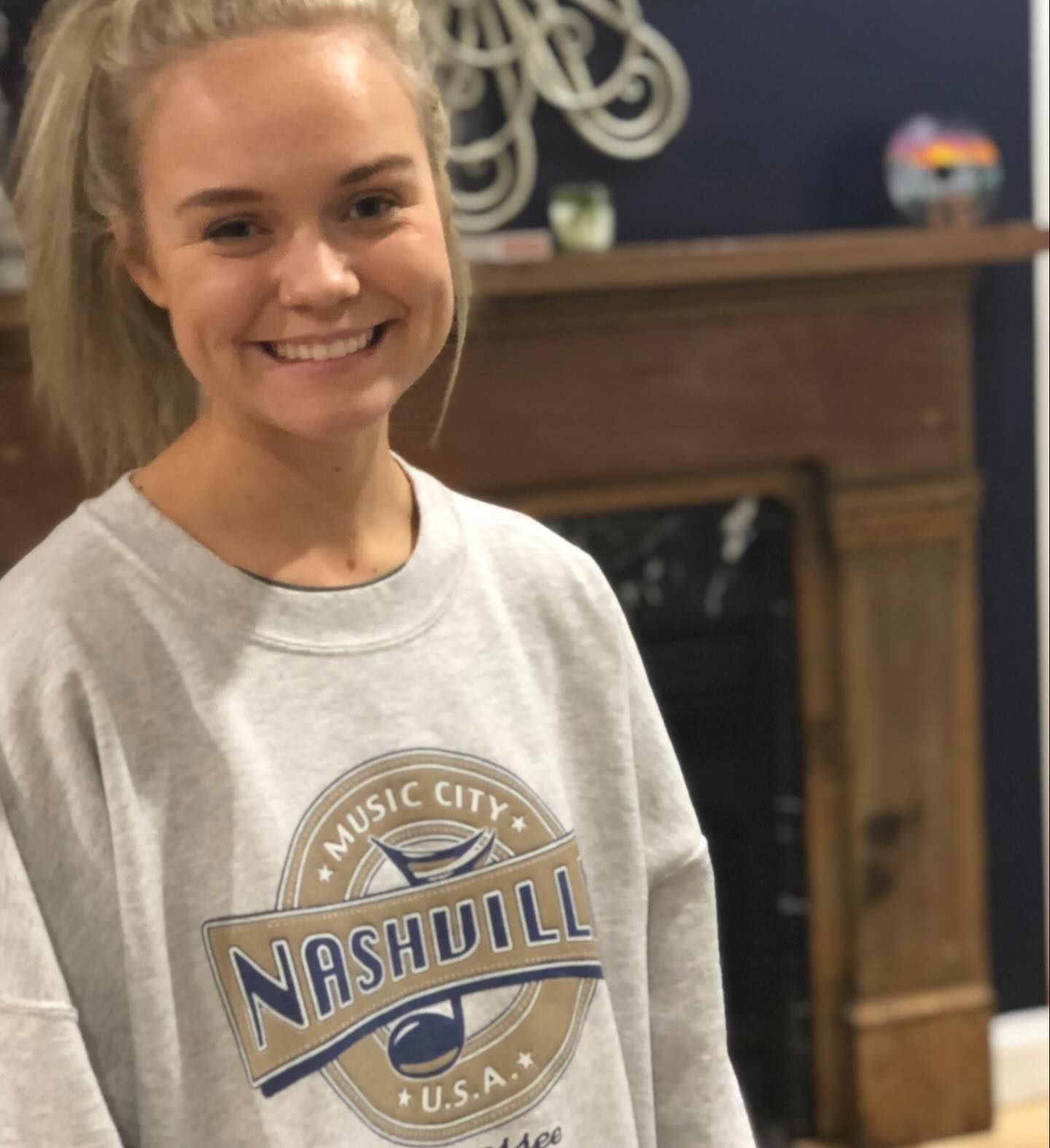 Young woman smiling, wearing a gray Nashville sweatshirt, posing near a fireplace, with blond hair up.