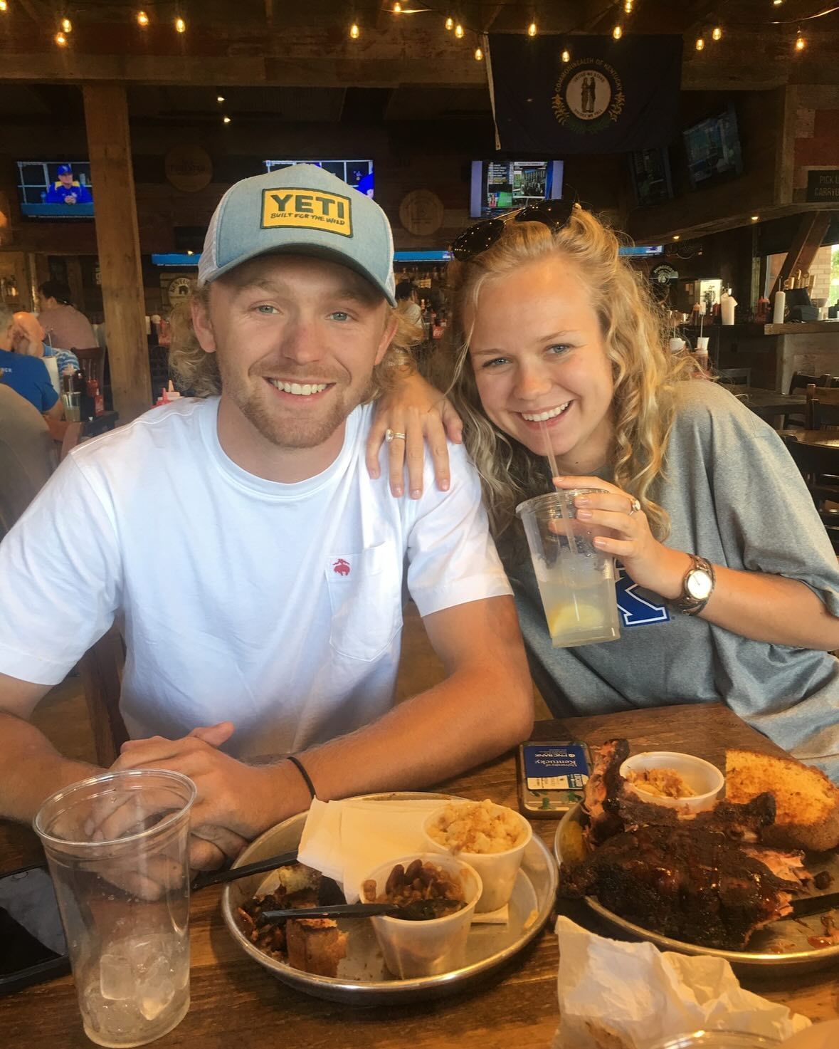 A smiling couple at a restaurant, plates of food in front of them. The man wears a Yeti hat, the woman holds a drink.