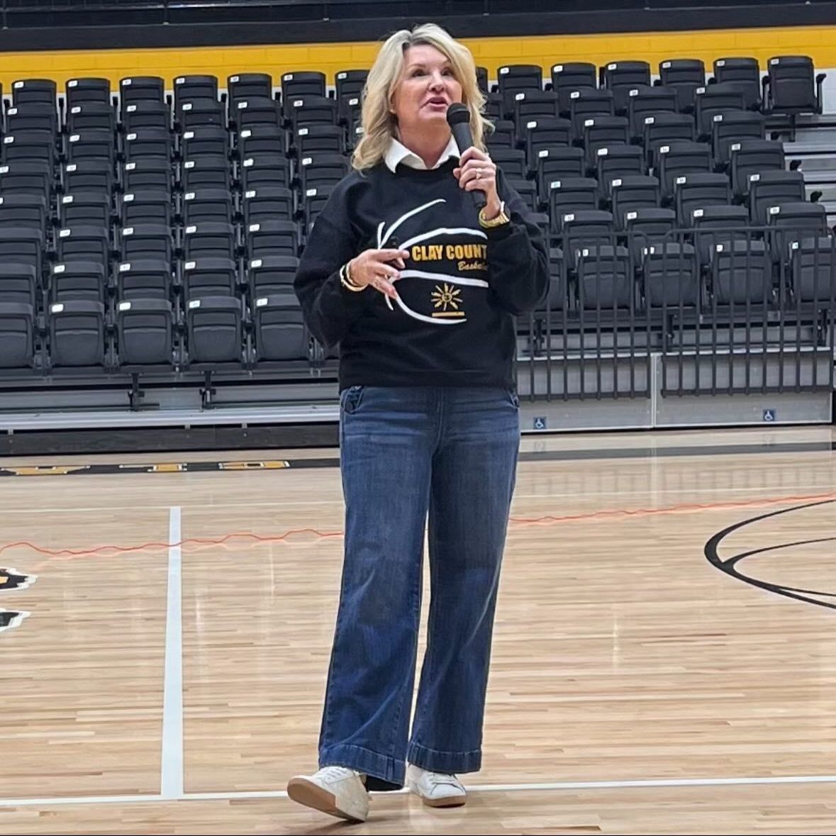 Woman speaking into a microphone on a basketball court. She wears a black sweatshirt, jeans, and white sneakers, with rows of empty bleacher seats in the background.