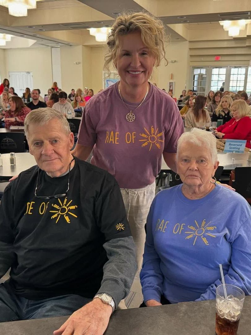 Three people smiling at a table. One in a purple shirt stands behind an elderly couple, all wearing shirts with a sun design. They are in a crowded cafeteria.