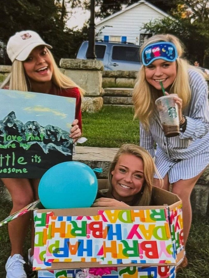 Three young women celebrating a birthday outdoors. One is in a decorated box, another holds a sign, and the other wears a sleep mask and sips a drink.