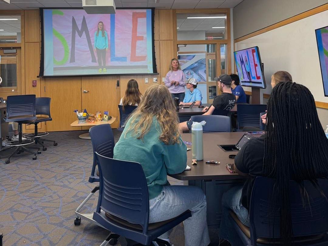 Students in a classroom listening to a presentation. A projection screen shows 