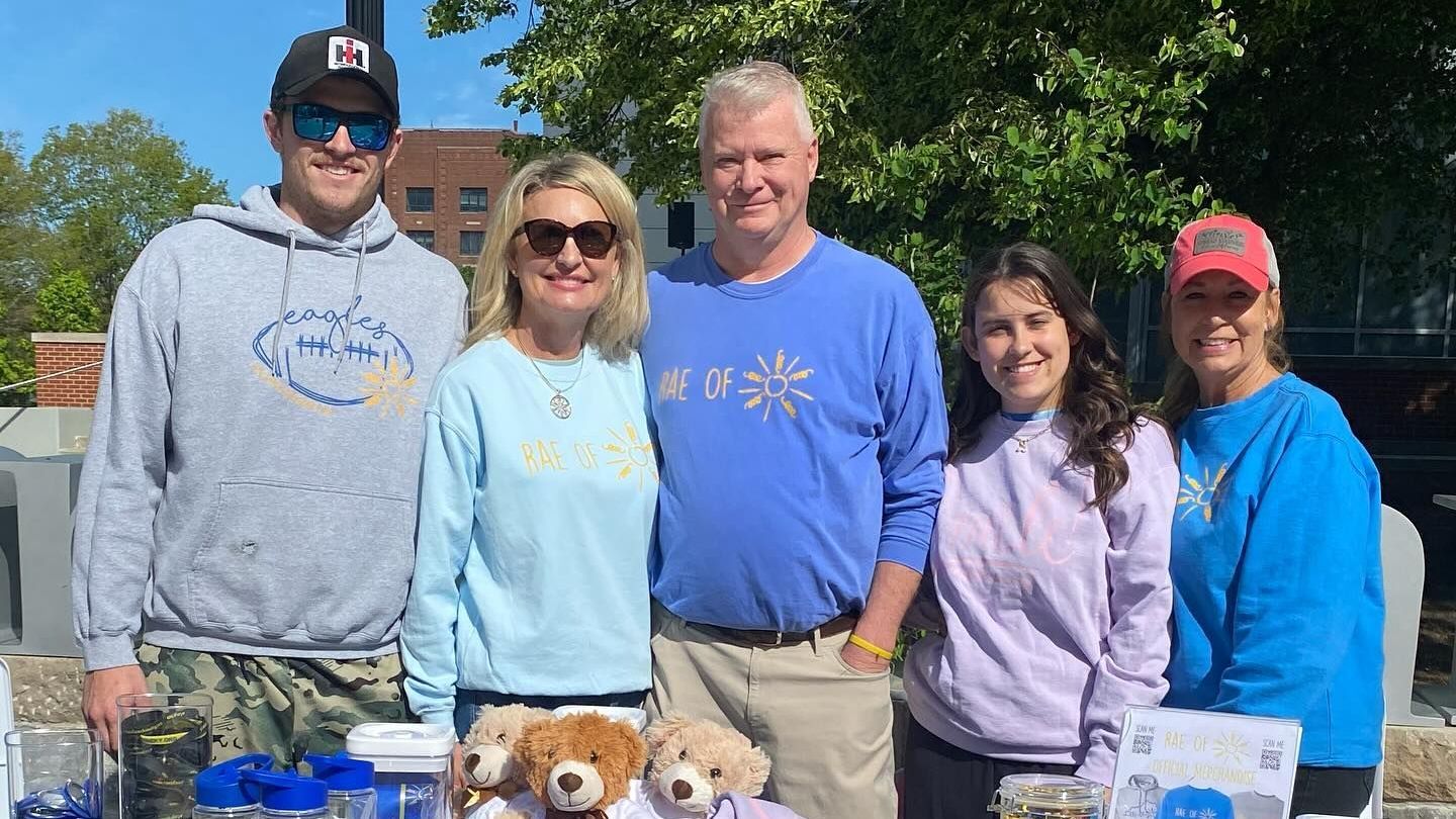 Five people stand behind a table outdoors, smiling and wearing blue and light-colored sweatshirts.