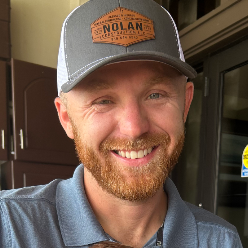 Man with reddish-brown beard and gray baseball cap smiles broadly. He's indoors, wearing a gray polo shirt.