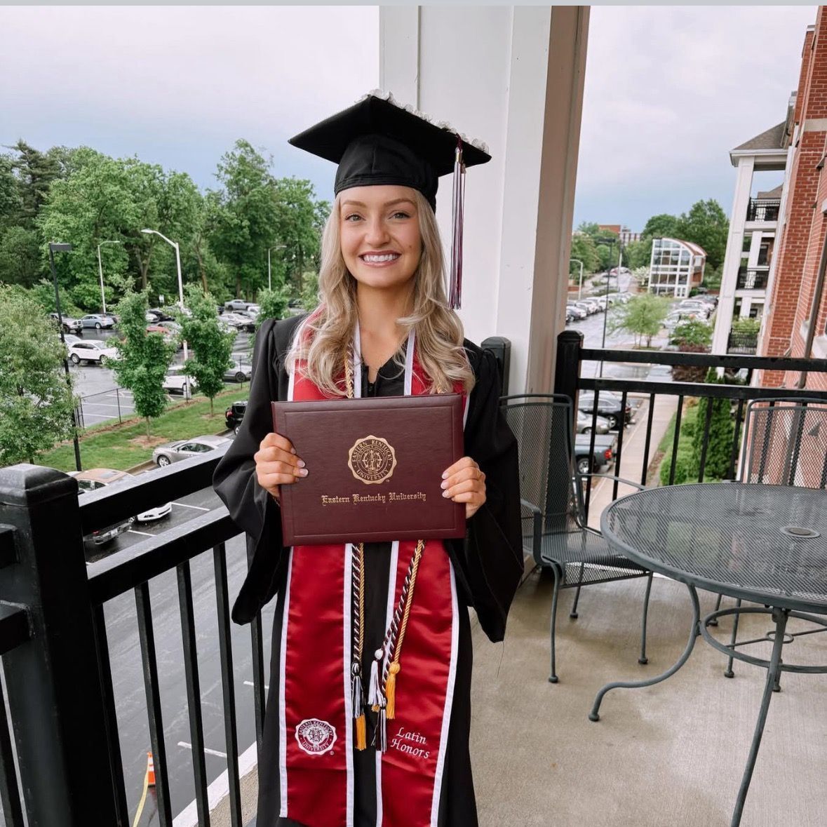 Woman in graduation attire stands on a balcony, holding diploma. She smiles, wearing a black gown, mortarboard, and red and white sash.