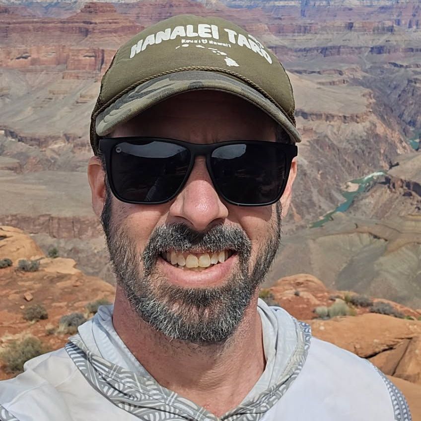Man with sunglasses and cap smiles at the camera, standing in front of the Grand Canyon.