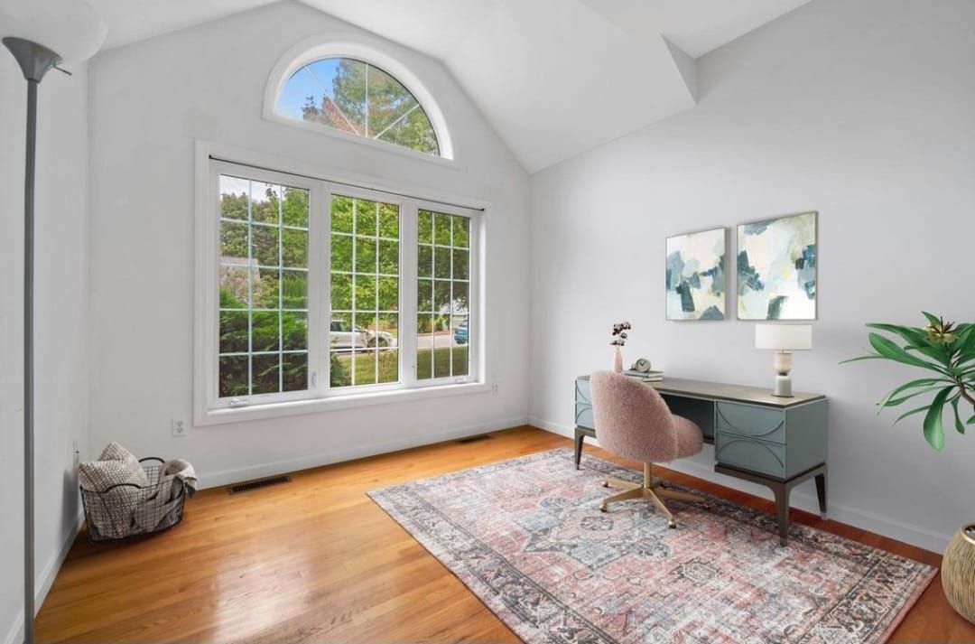 Home office with desk, large window, rug, and pink chair. White walls and hardwood floors.