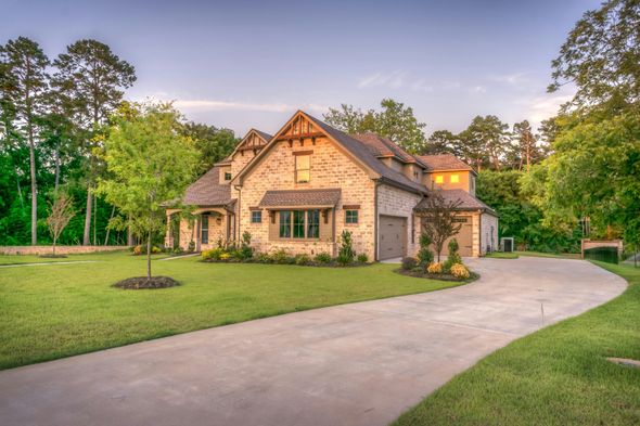 Brick house with a long concrete driveway, green lawn, and surrounding trees in a well-lit setting.