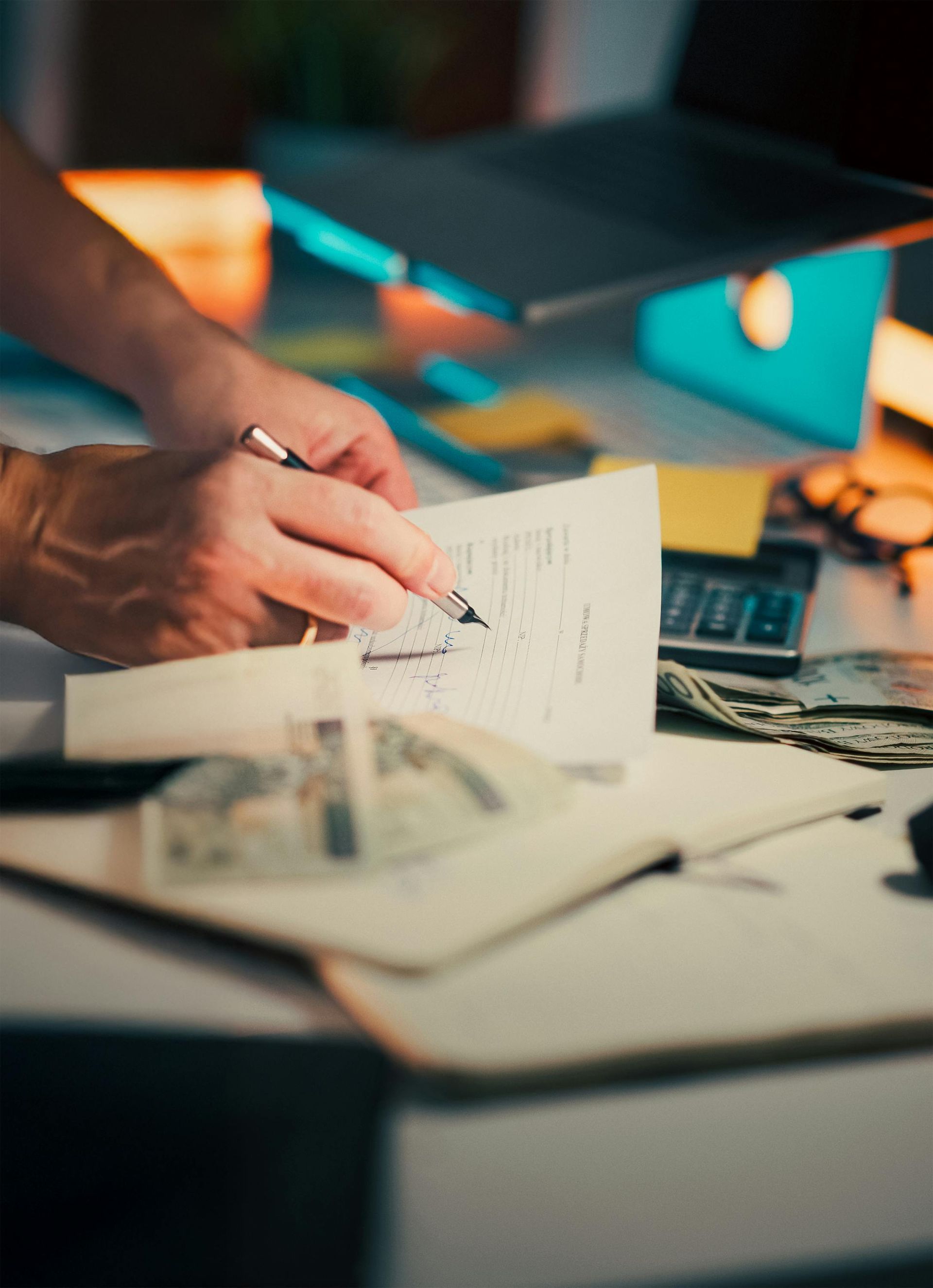 Person's hands writing on a document at a desk with money, a calculator, and a laptop.